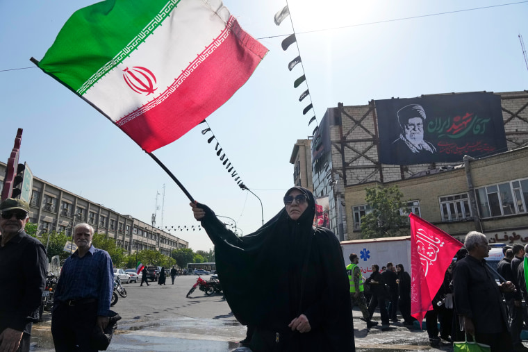 Image: A woman waves an Iranian flag. A banner showing a portrait of the late Supreme Leader Ayatollah Ali Khamenei is seen hanging on a building wall in the background.