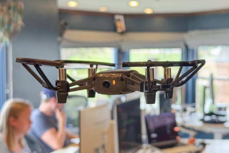 A drone flies in the air inside of a home, people can be seen working on computers at desks in the background