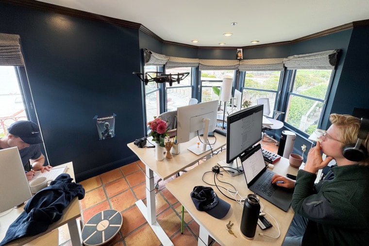 People work inside of a home office at desks with computers on them, a drone flies in the air off from a circular drone pad seen on the floow