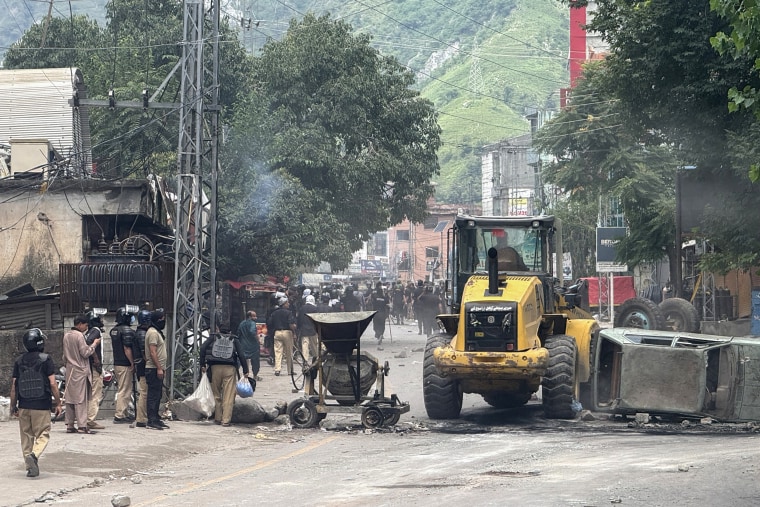 Police personnel watch as workers clear a road blocked by protesters and debris.