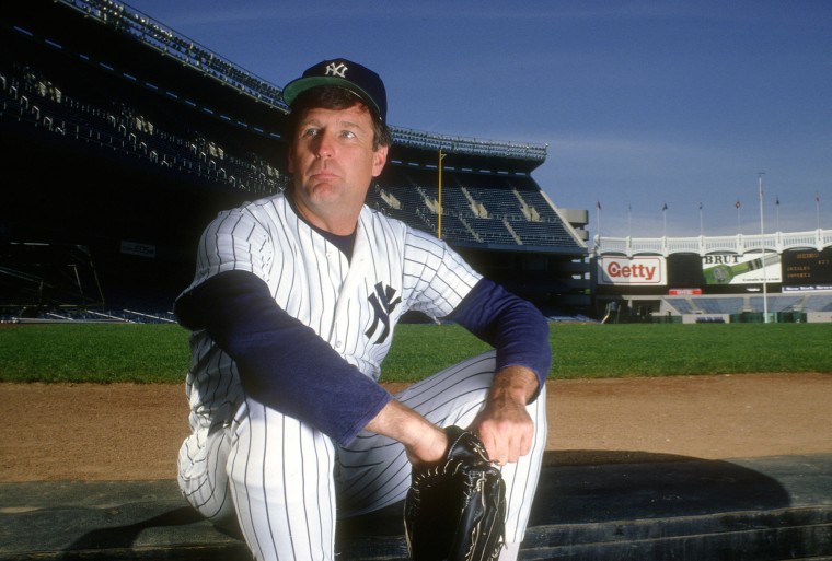 Tommy John sits on the baseball field in uniform, holding a catcher's mitt