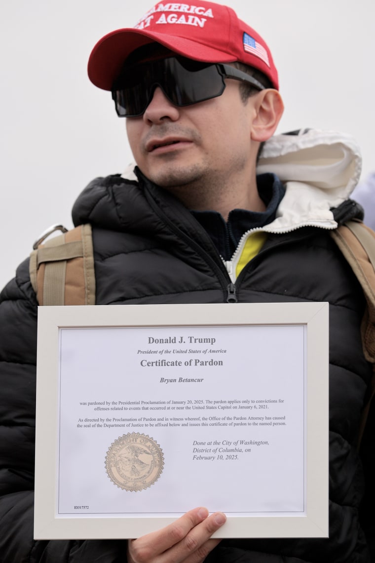 A man wearing a Make America Great Again hat and dark sunglasses holds a framed certificate of pardon from President Trump.