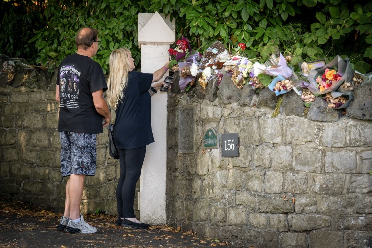 A couple placing a bouquet of flowers among others stacked on a stone wall.