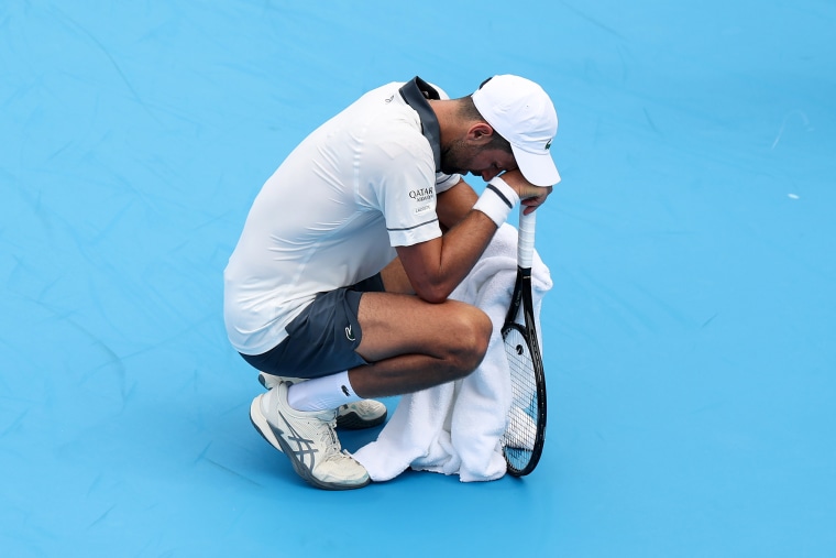 Novak Djokovic squats down and rests his head on the tennis racket he's holding, while also holding a towel