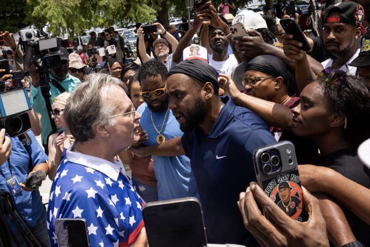 Two men, one wearing a shirt decorated like the American flag, confront each other during an outdoor demonstration.