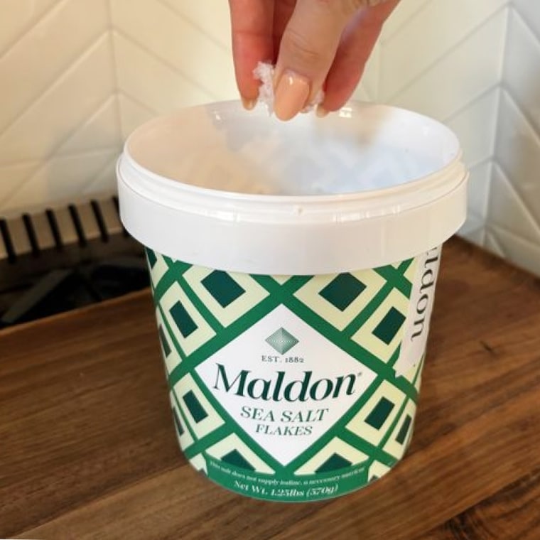 A hand reaching for a pinch of Maldon Sea Salt Flakes in the 20-ounce tub.
