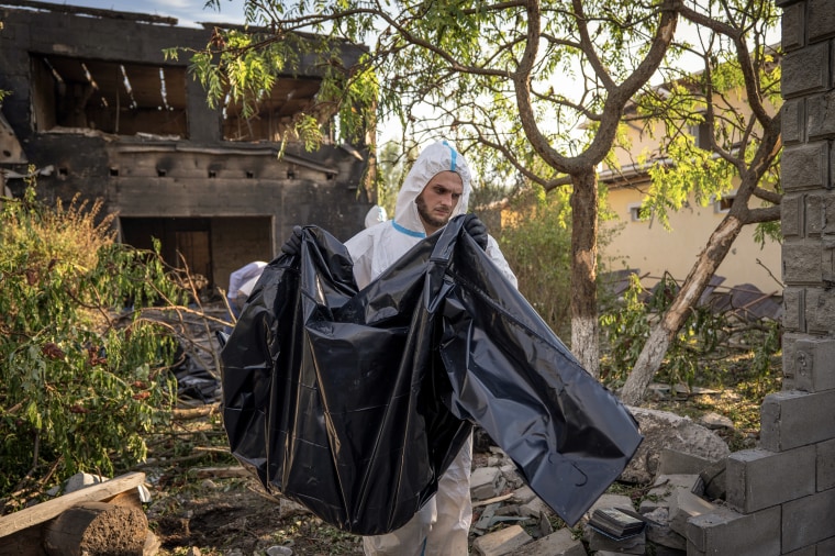 A man wearing white coveralls carries a body wrapped in black plastic sheeting.