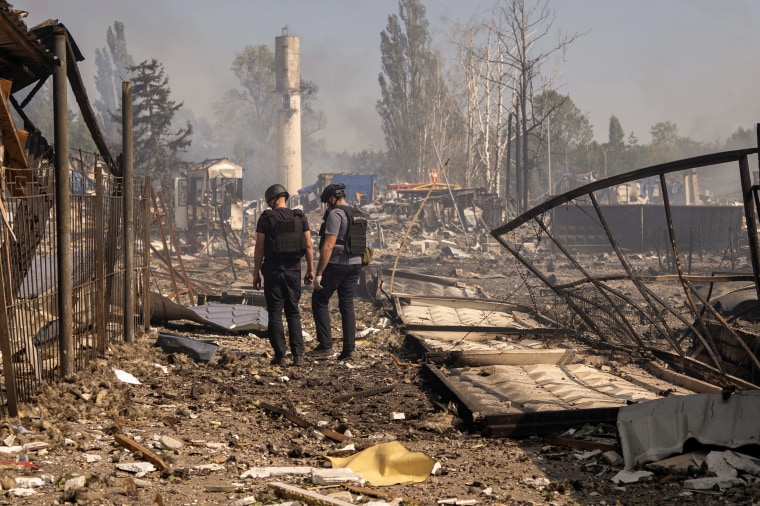 Two men wearing helmets and flak jackets stand amidst extensive damage following an explosion.