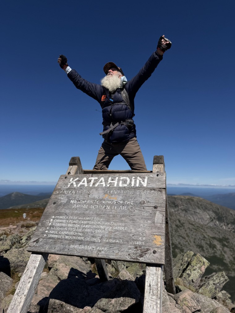 Dale Sanders celebrates with wide open arms on top of a hike sign at the summit of Mt. Katahdin.