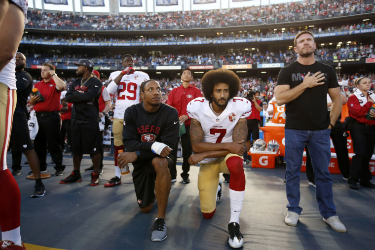 Reid and Kaepernick take a knee on the sidelines of a football stadium while another player holds his hand over his heart.