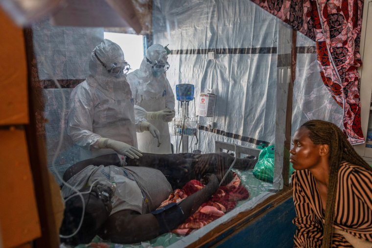 Health workers tend to a patient while a woman observes from the right.
