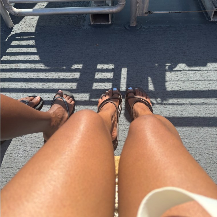 Two feet wearing the Crocs Miami sandals in brown beside two feet wearing plain black flip flops with a grey background.