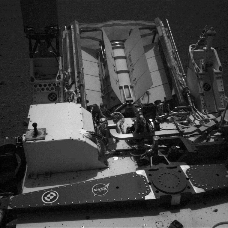 A view from the Navcam system on the Curiosity rover's camera mast looks toward the back of the rover. The most prominent hardware is the probe's radioisotope thermal generator, or RTG, which uses radioactive plutonium to generate electricity. The fins serve as radiators for heat from the RTG. The picture also shows black bits of Martian debris that were thrown onto the rover's deck during landing. The original picture has been processed to enhance brightness.