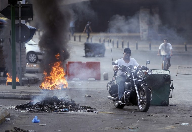 A man rides his motorcycle between a burning tire and garbage containers laid by Sunni protesters after overnight clashes between Sunni and Shiite gunmen in Beirut, Oct. 22, 2012.