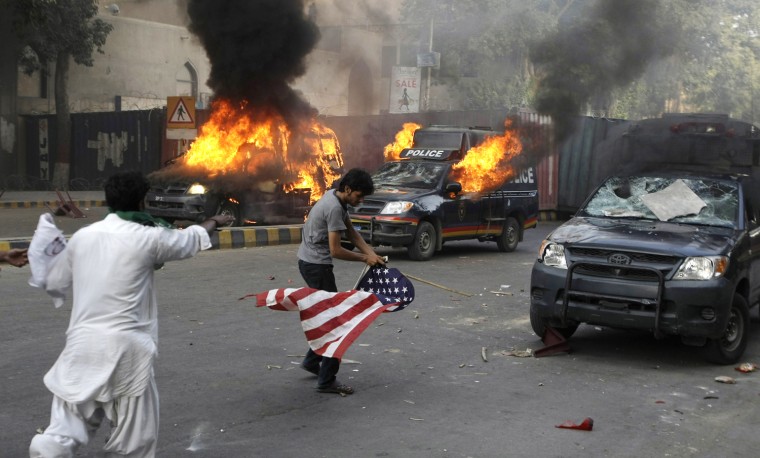 A protester carries a representation of a U.S. flag as police vehicles burn in Karachi, Pakistan on Sept 21.