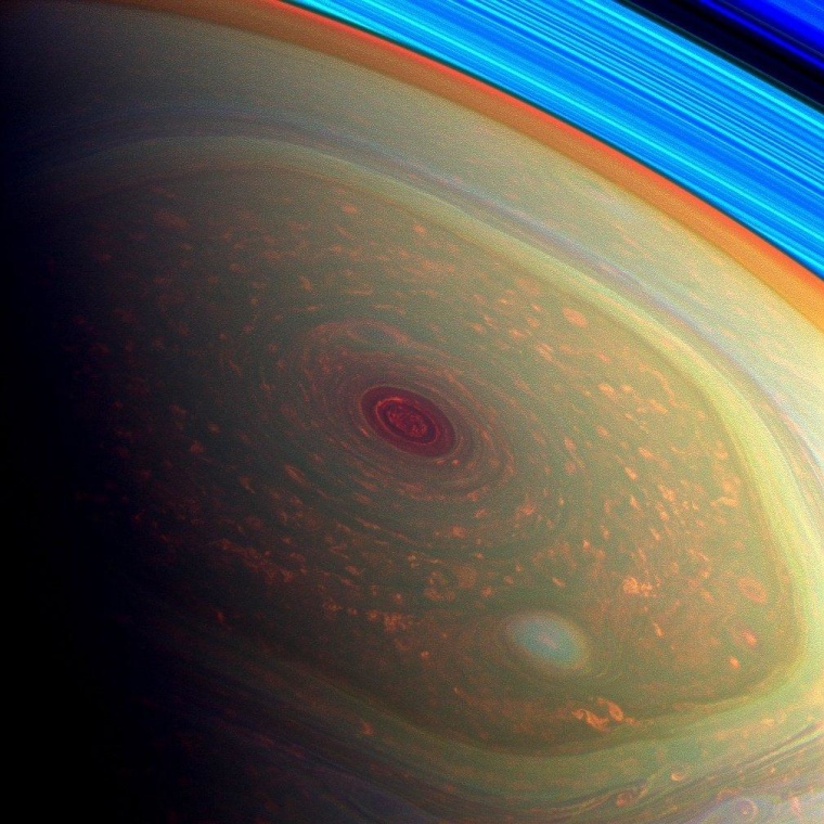 A false-color image from NASA's Cassini mission highlights the storms at Saturn's north pole. The angry eye of a hurricane-like storm appears dark red, while the fast-moving hexagonal jet stream framing it is a yellowish green. Low-lying clouds circling inside the hexagonal feature appear in a muted orange color. A second, smaller vortex pops out in teal at the lower right of the image. The rings of Saturn appear in vivid blue at the top right. The colors are coded to show different near-infrared wavelengths, which correlate to different altitudes in the planet's polar atmosphere.
