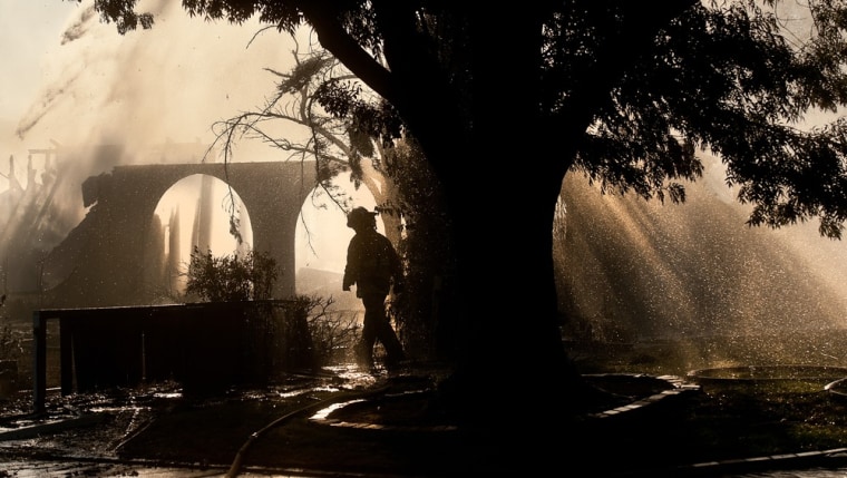 As water sprays from a hose above, a firefighter passes one of several Marigold Drive homes scorched after a grass fire spread to structures.