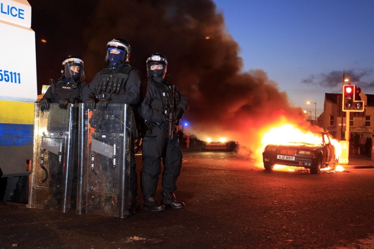 Police officers in riot gear stand near a burning, hijacked car during rioting in East Belfast on Saturday. Protests continue in Northern Ireland as loyalists renewed their anger against restrictions on flying the union flag from Belfast City Hall.