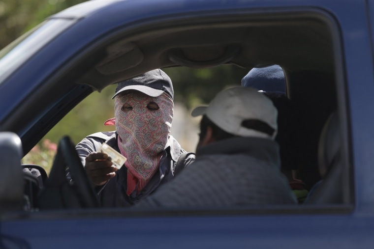In this photo taken Friday, a masked and armed man checks the identity of a driver at a roadblock at the entrance to the town of El Pericon, Mexico.