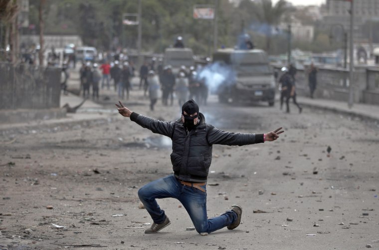 A masked Egyptian protester flashes the victory sign during clashes with police, background, near Tahrir Square, Cairo, on Jan. 28.