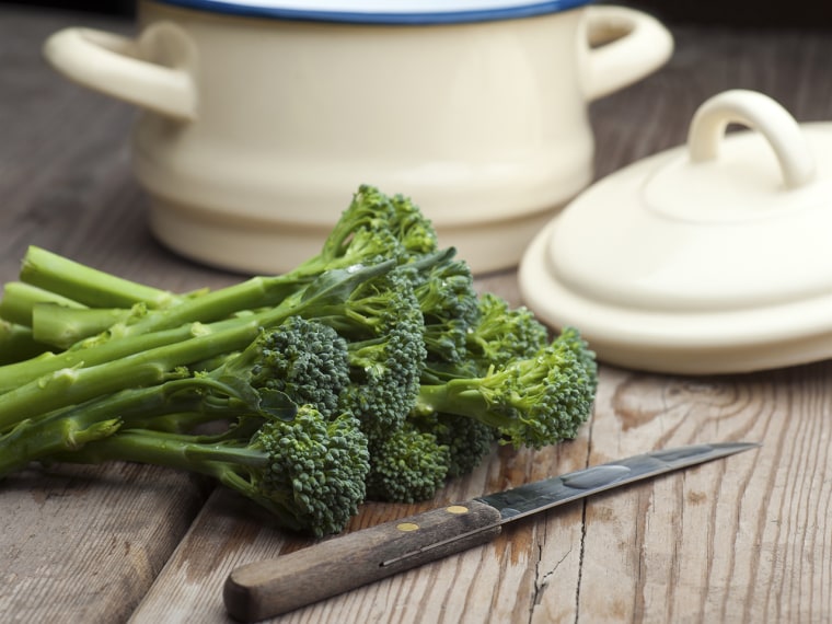 Fresh Broccoli Laid On A Wooden Kitchen Table With A Vegetable Knife and A Pan With Lid In The Background