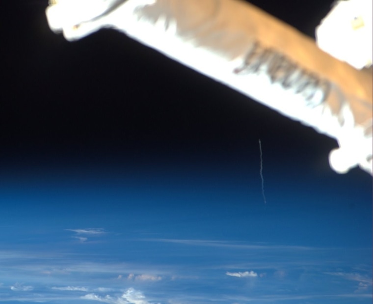 This view from the International Space Station documents the launch of the European Space Agency's Kepler cargo spaceship as a wisp of smoke toward the right side of the image. A section of the station's robotic arm dominates the foreground.