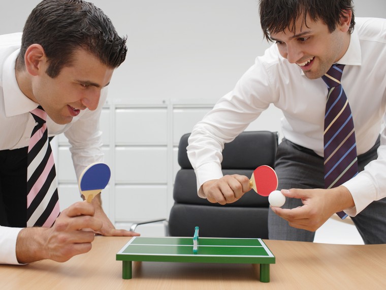 Businessmen playing minature table tennis at desk.