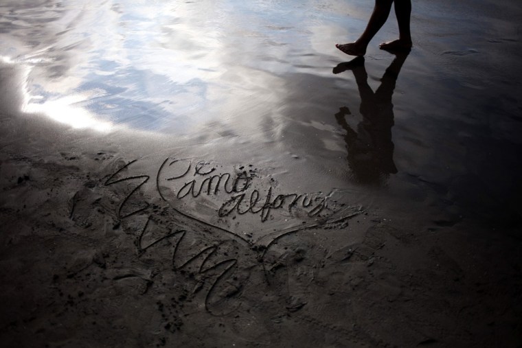 A man walks along the shoreline next to a message in the sand that reads in Spanish