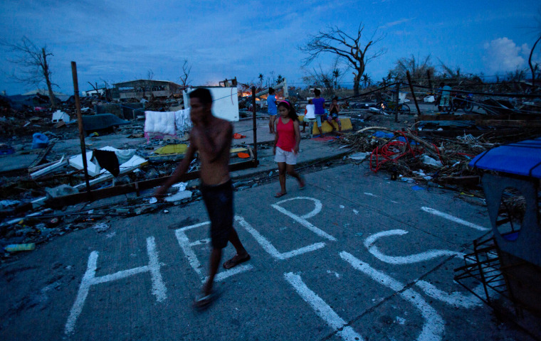 Typhoon Haiyan survivors walk through the ruins of their neighborhood in Tacloban, Nov. 13, 2013, where a man named J.R. Apan painted a plea for help in front of his destroyed home the day after the typhoon hit hoping for aid to arrive but says he has not yet received food or water supplies.