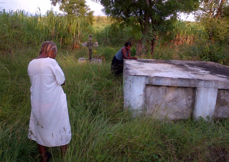 Lisette Paul (leaning on crypt) and her mother Dieumene Bastien say that some family members who died of cholera were put in plastic bags and buried in holes in the ground because people were afraid of contracting the disease during a proper burial. One cholera victim, Dieumene's son – Lisette's brother – is buried in the crypt.