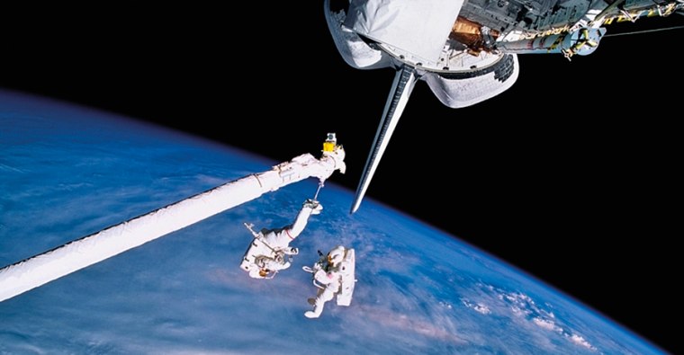 Astronauts Carl J. Meade and Mark C. Lee (red stripes on spacesuit) work in space while testing the SAFER backpack system (Simplified Aid For EVA Rescue) in September 1994. Astronaut Lee can be seen attached to Discovery's Remote Manipulator System robotic arm.