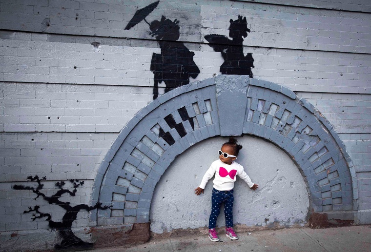 A child poses for a photo under a piece by British graffiti artist Banksy in the Brooklyn borough of New York, Oct. 17, 2013.