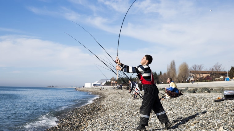 Men cast their fishing rods at the Black Sea beach with the Fisht Stadium (rear) in the Olympic Park visible in the background, Sochi, Rus...