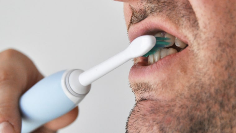 Man brushing his teeth with electric toothbrush, caucasian male with beard in front of solid coloured light background stock image