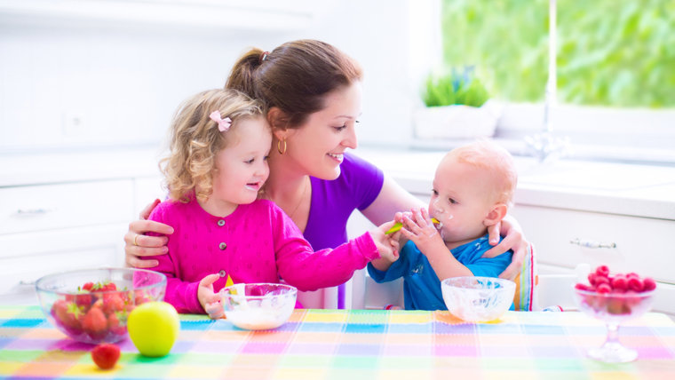 Happy young family, mother with two children, adorable toddler girl and funny baby boy having healthy breakfast eating fruit and dairy, sitting in a w...