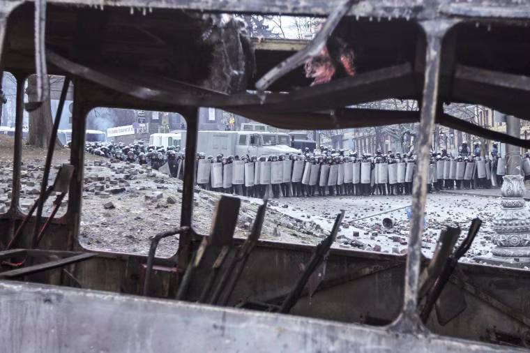 Image: Ukrainian riot police take cover behind their shields in front of a burnt bus during a rally near government administration buildings in Kiev