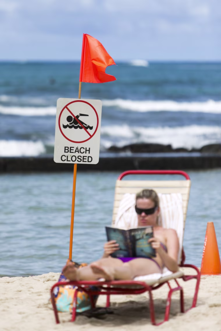 Image: A sunbather reads a book behind a beach closed sign at Waikiki Beach in Honolulu, Hawaii