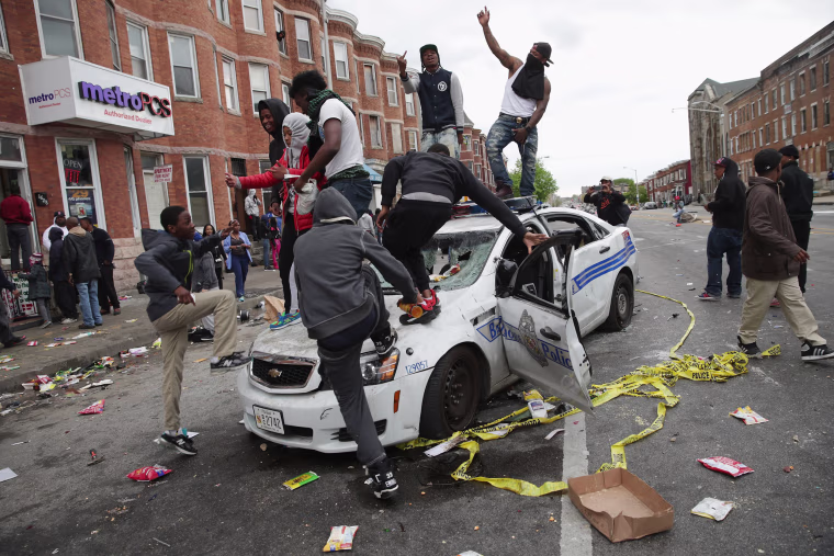 Image: Demonstrators climb on a destroyed Baltimore Police car in the street