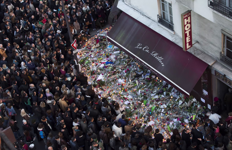 Image: A large crowd gathers to lay flowers and candles in front of the Carillon restaurant in Paris