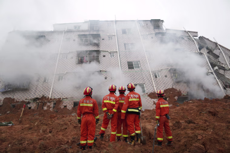 Image: Firefighters look on as smoke rises in front of a damaged building at the site of a landslide at an industrial park in Shenzhen