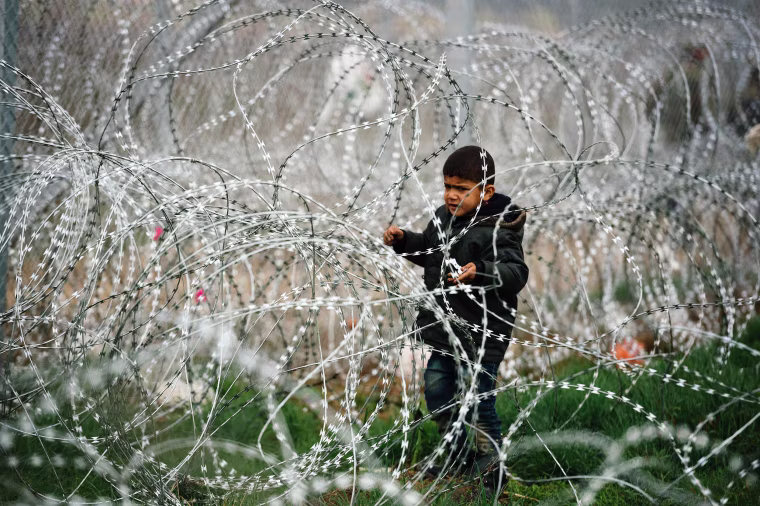 Image: A child stands among a razor-topped fence close to the gate at the Greek-Macedonian border