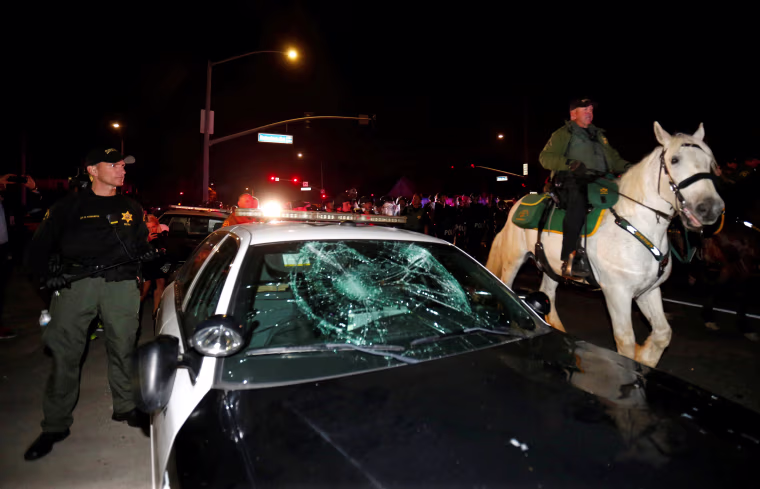 Image: Police on horseback and in riot gear pass by a damaged police car as they break up a demonstration outside Republican U.S. presidential candidate Donald Trump's campaign rally in Costa Mesa