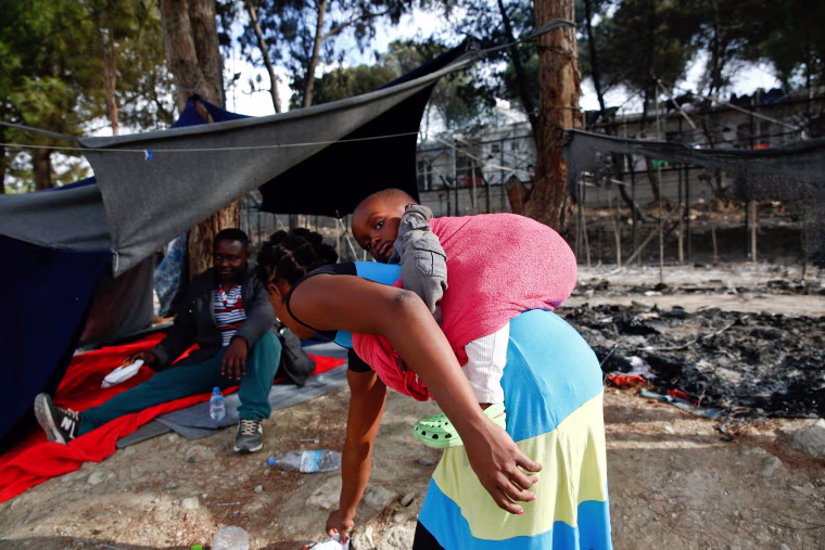 Image: A migrant woman carries a baby as she stands next to the remains of a burned tent at the Moria migrant camp