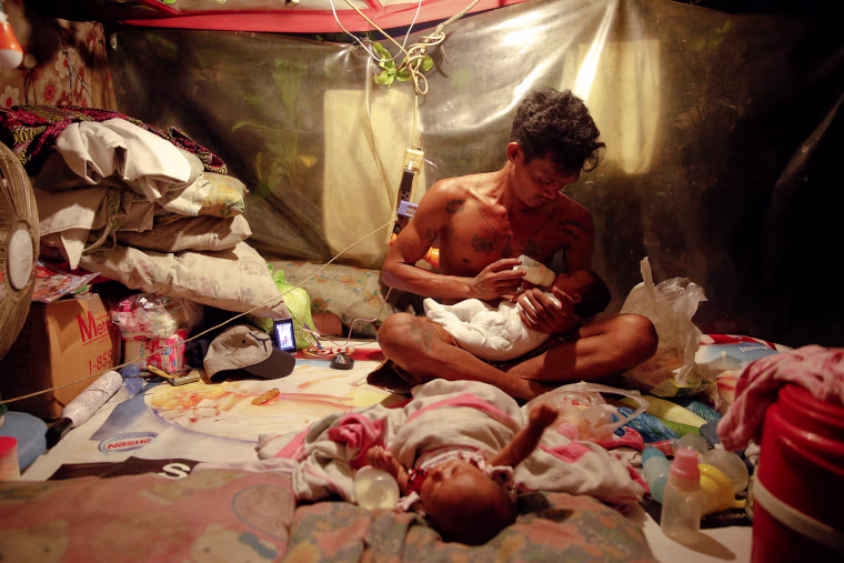 Image: A man who lives with his family in a tent erected atop a tombstone, feeds his two-month old twins at the North Cemetery where many victims of country's war on drugs are buried in Manila