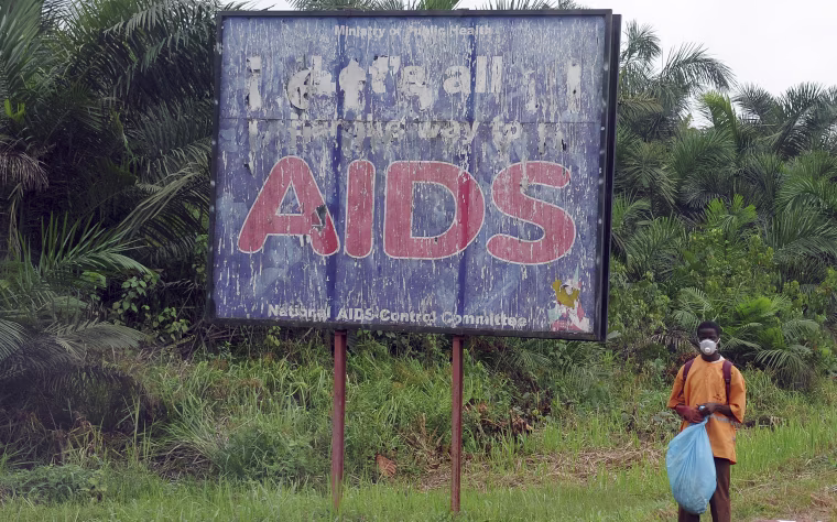 Image: A man stands next to an AIDS billboard as he cleans a street in Yaounde