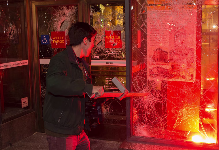 Image: A man pulls a piece of metal out of a window as a flare burns inside a Wells Fargo Bank
