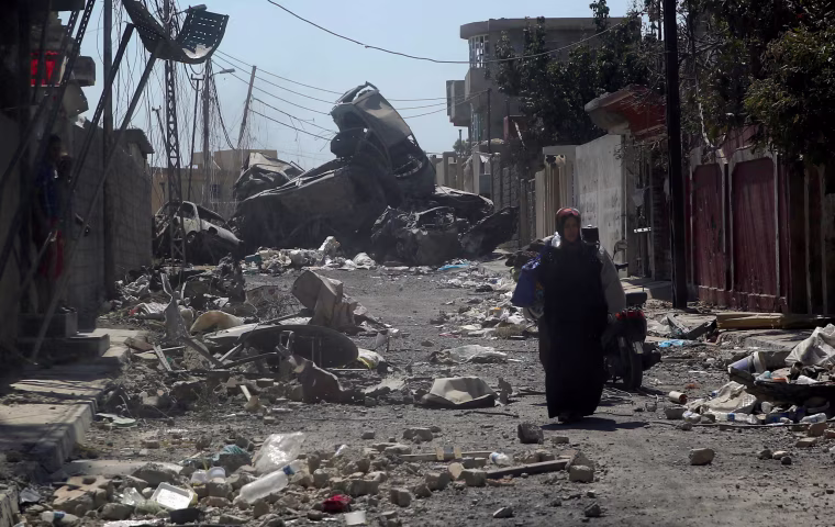Image: Displaced Iraqi civilians walking along a road littered with debris and destroyed vehicles are pictured behind a car glass window in western Mosul