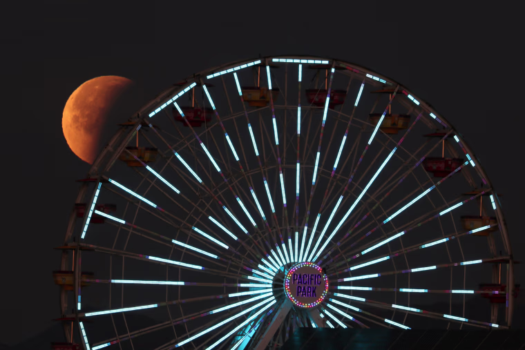Image: A lunar eclipse of a full \"Blue Moon\" is seen above the ferris wheel on the Santa Monica Pier in Santa Monica