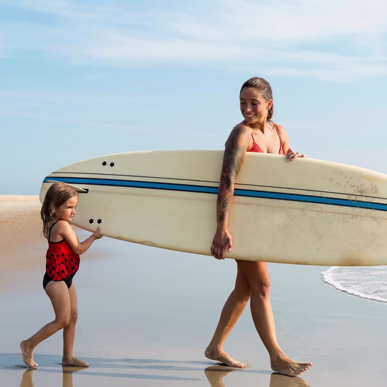 Mother and daughter walking on beach holding surfboard