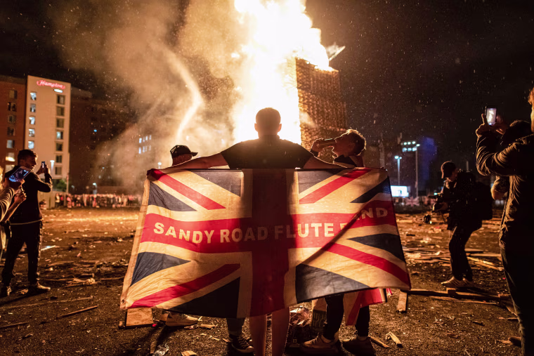 Image: A man drapes the Union Jack around his shoulders in front of a blazing pyre at the Sandy Row bonfire build in South Belfast.
